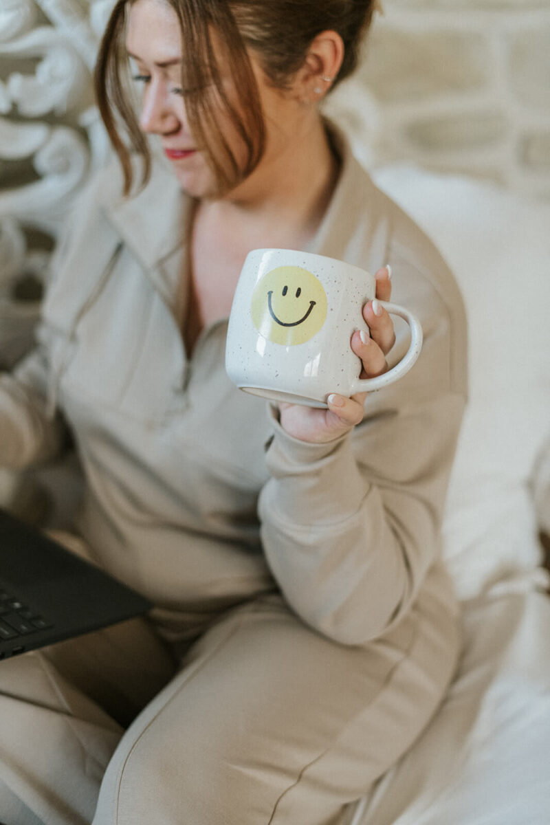 Angee with hair in ponytail working and holding a coffee mug with a smiley face on it.