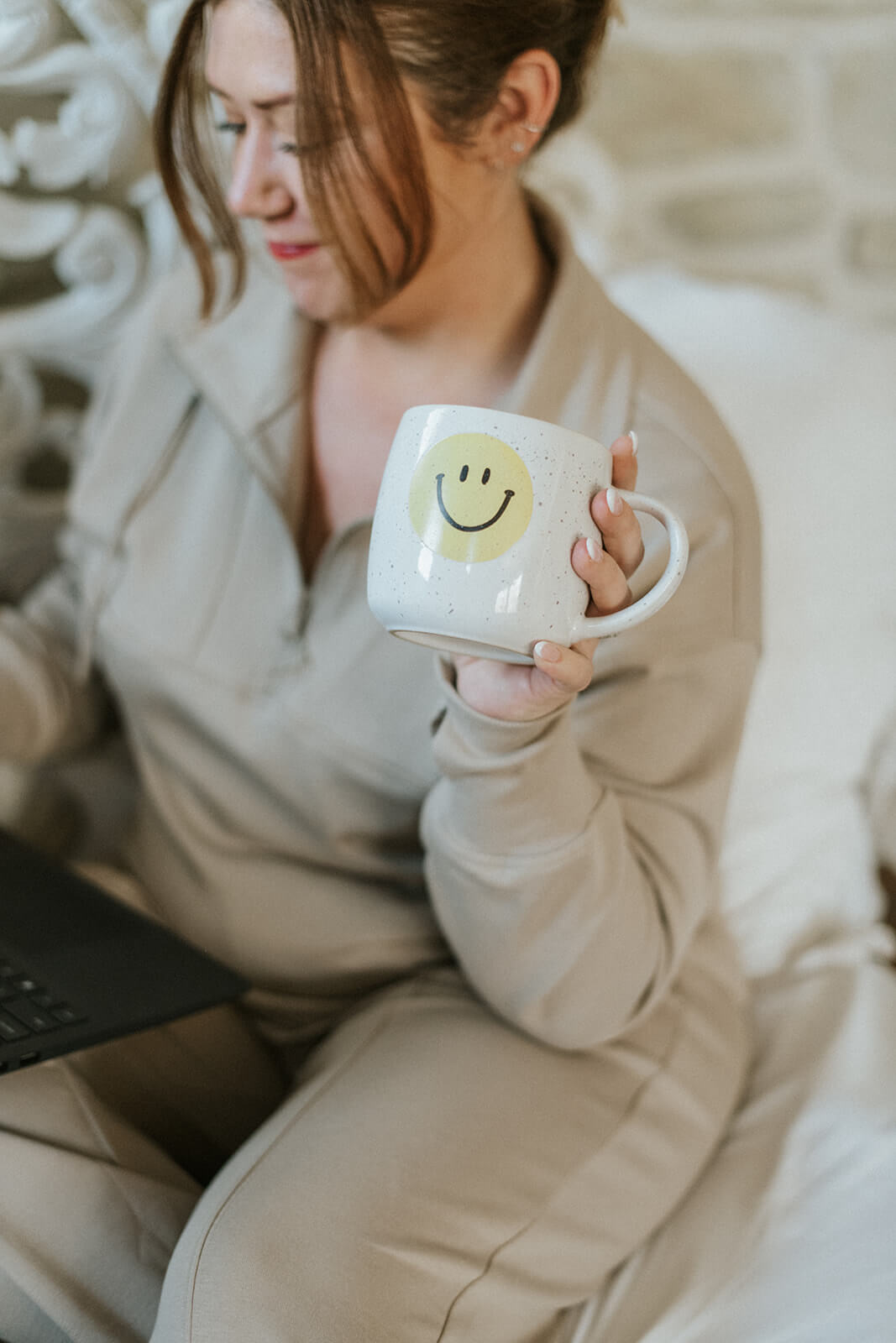Angee with hair in ponytail working and holding a coffee mug with a smiley face on it.